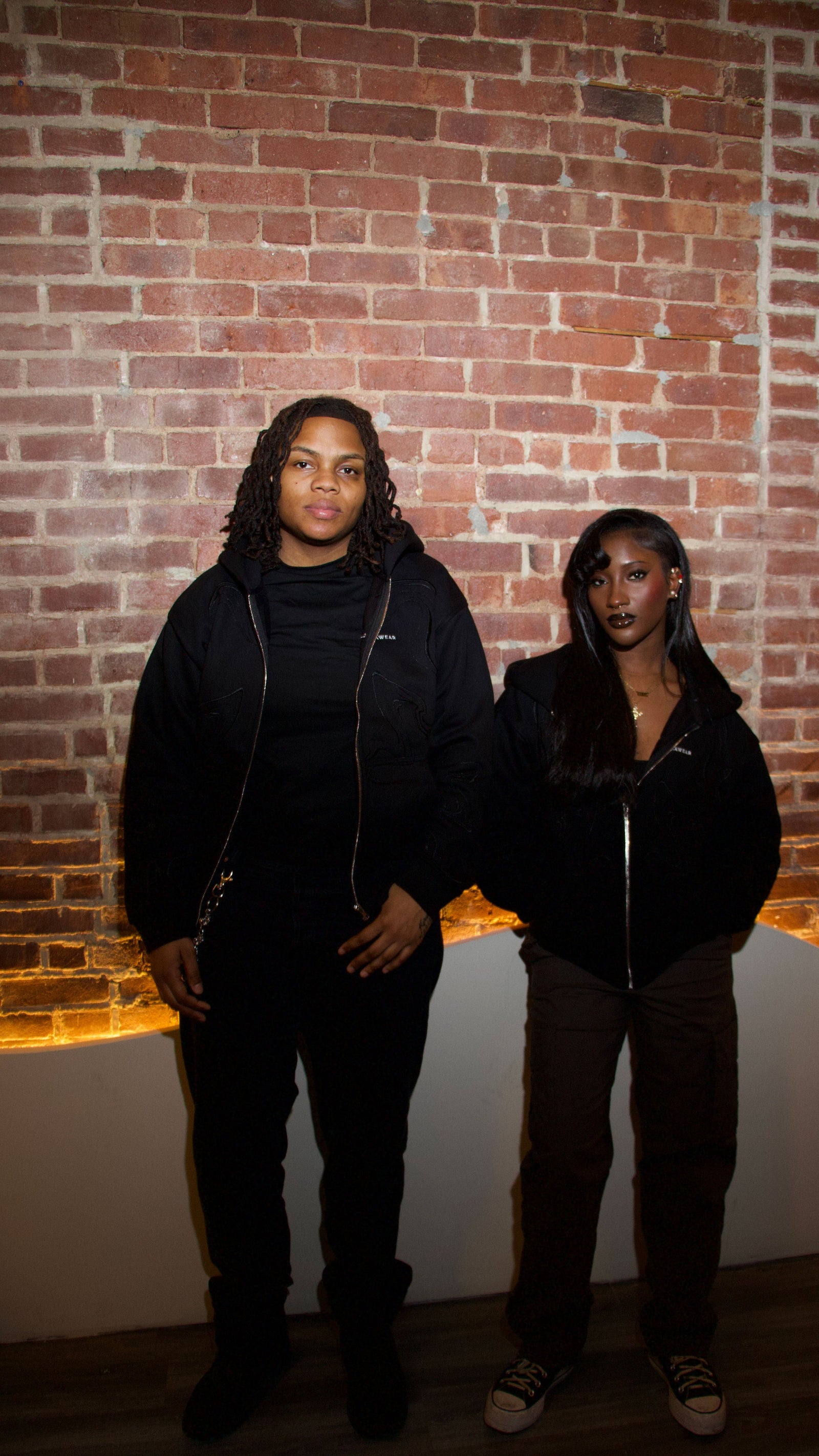 Two people standing against a brick wall with warm lighting